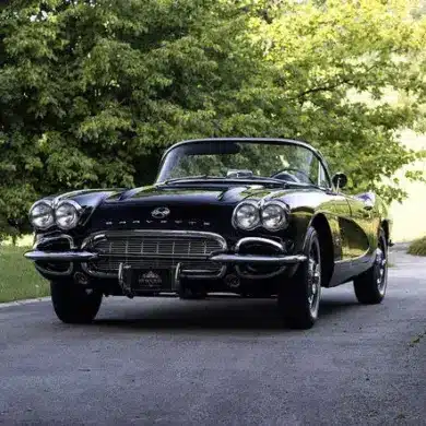 Eine schwarze Chevrolet Corvette C1 Cabriolet von 1961 steht auf einer asphaltierten Allee, umgeben von grünen Bäumen.