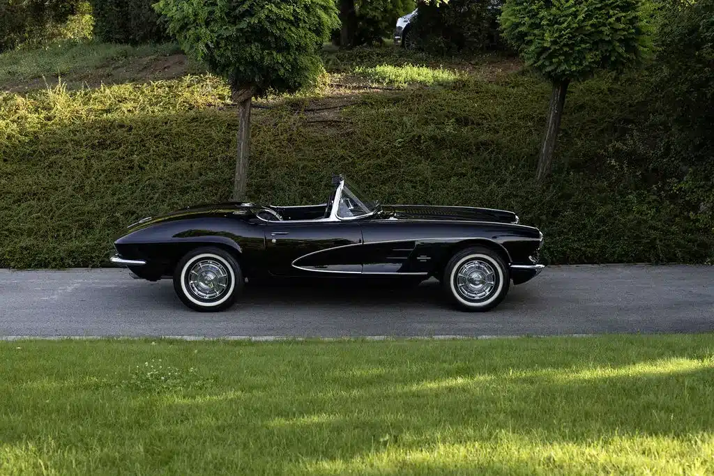 Schwarze Chevrolet Corvette C1 Cabriolet von 1961, restauriert, Seitenansicht auf einer Asphaltstraße vor grüner Hecke.