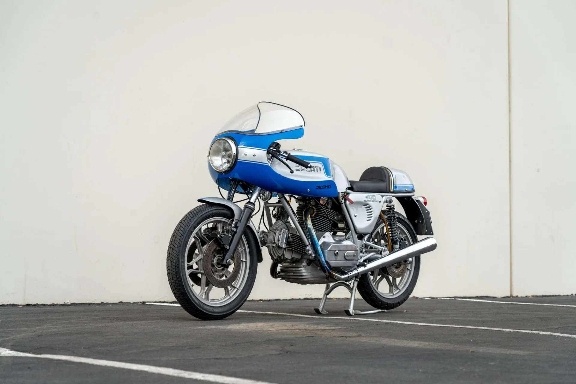Seitenansicht einer Ducati 900 Super Sport von 1975 in Blau und Silber, stehend auf einem Parkplatz vor einer hellen Wand.