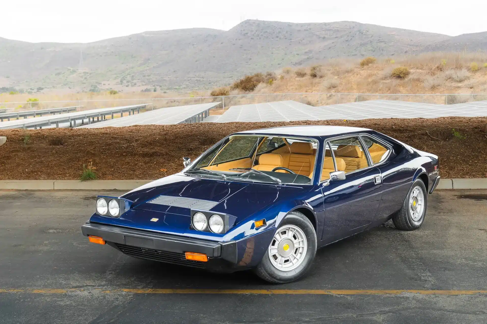 Blauer Ferrari Dino 308 GT4 von 1975 mit beigefarbenem Interieur auf einem Parkplatz vor einer Hügellandschaft