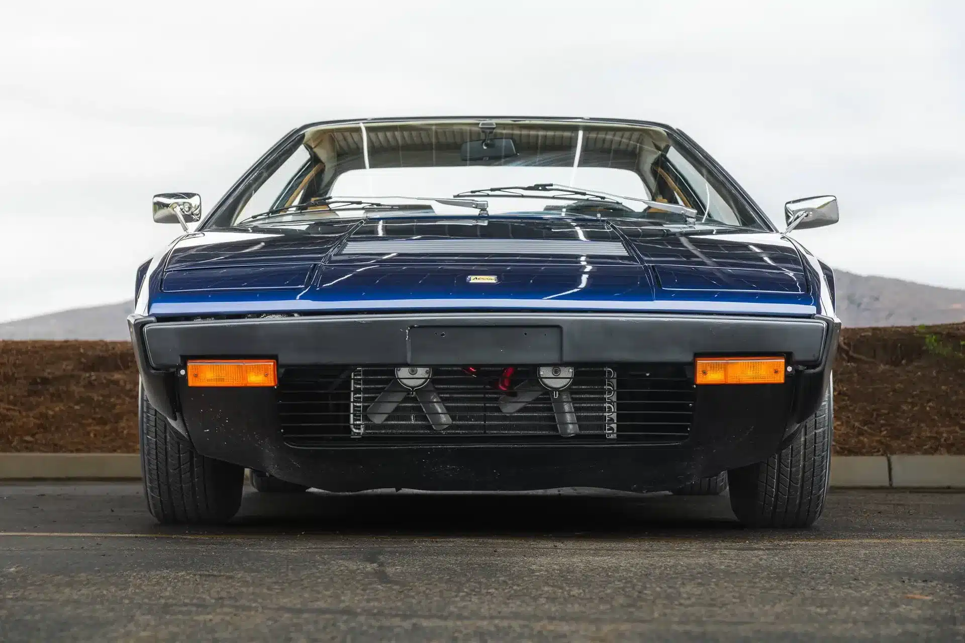 Frontansicht eines restaurierten Ferrari Dino 308 GT4 von 1975 in blauer Lackierung, aufgenommen auf einem Parkplatz.