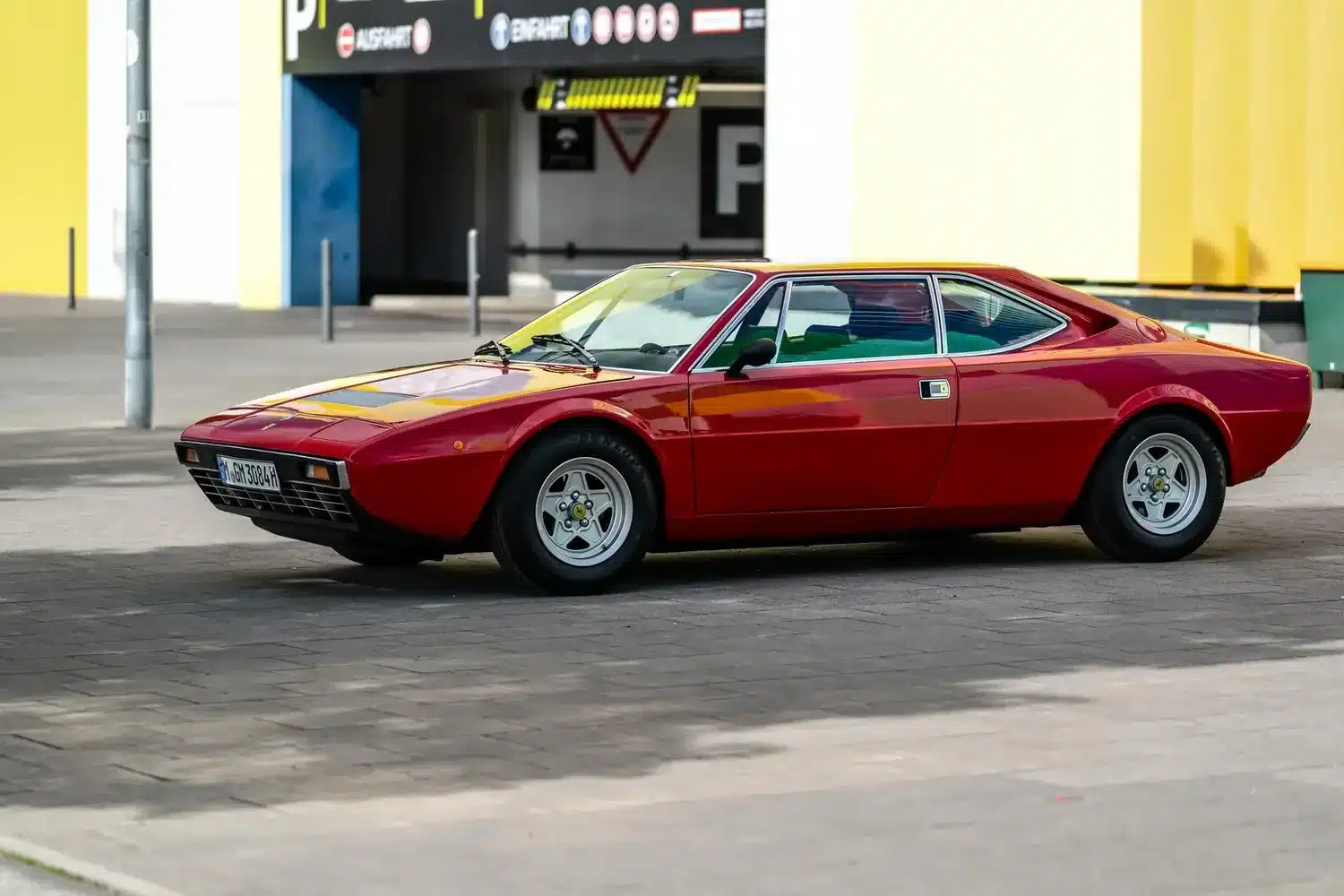 Roter Ferrari Dino 308 GT4 Baujahr 1975 mit kantigem Design, geparkt seitlich vor Parkhaus.