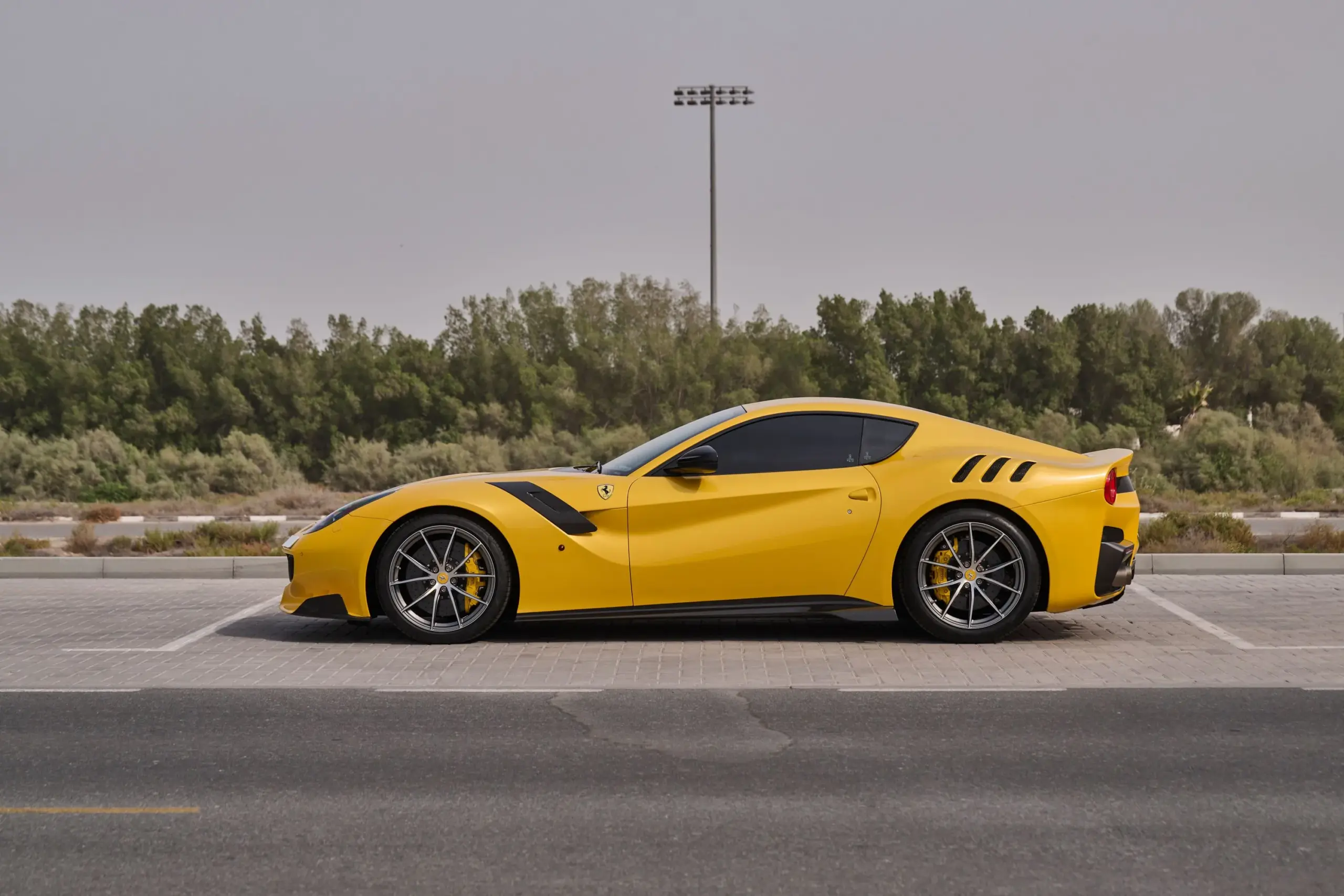Ferrari F12 TDF in gelber Lackierung mit schwarzen Carbon-Details, Seitenansicht auf Parkplatz, Bäume im Hintergrund.