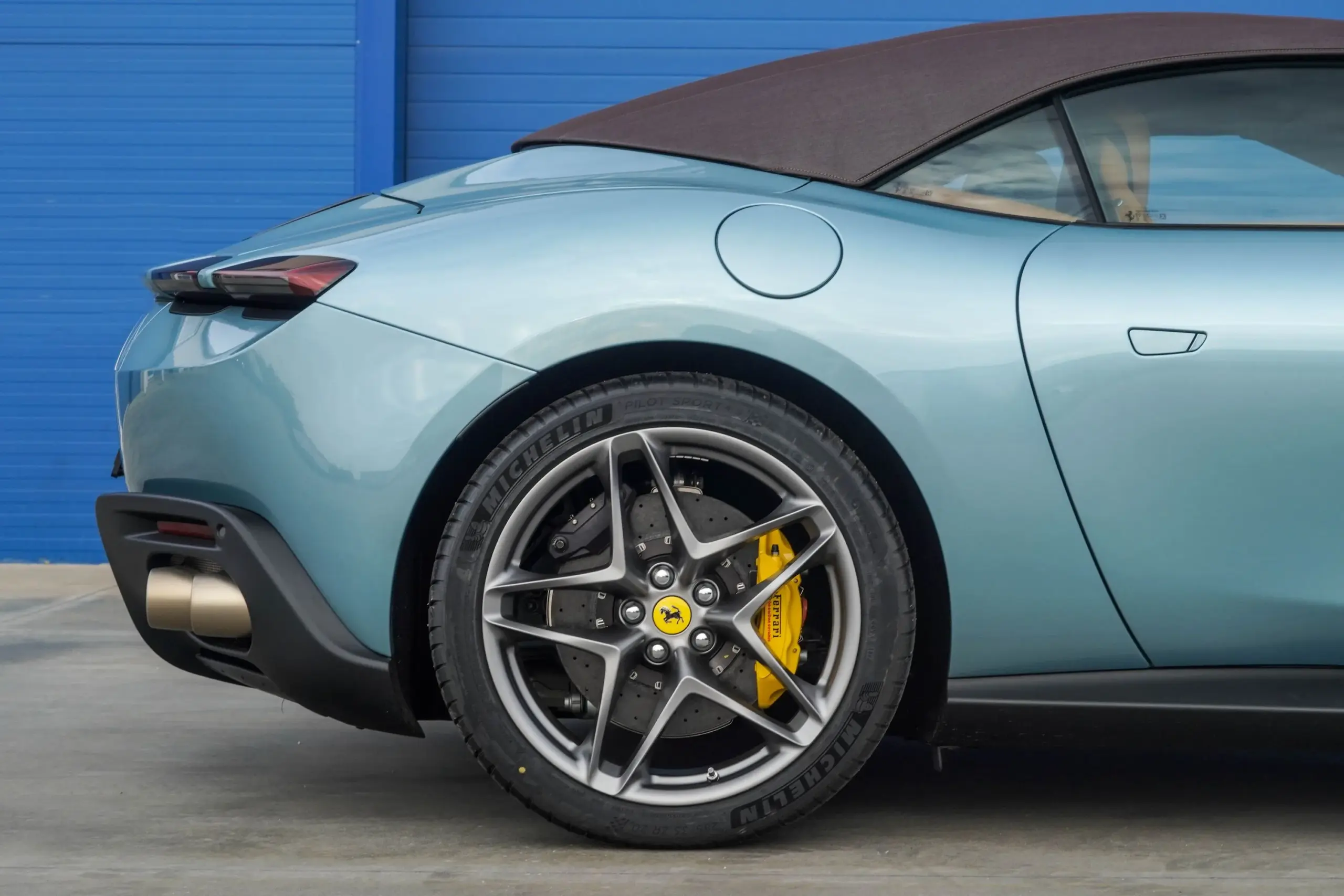 Seitliche Heckansicht eines Ferrari Roma Spider 2025 in Celeste Trevi mit dunkelbraunem Stoffverdeck und matten 20-Zoll-Schmiedefelgen.