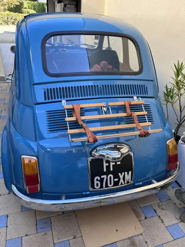 Blauer Fiat 500L von 1968 von hinten mit offenem Faltdach und Holzgepäckträger, aufgenommen im Außenbereich.