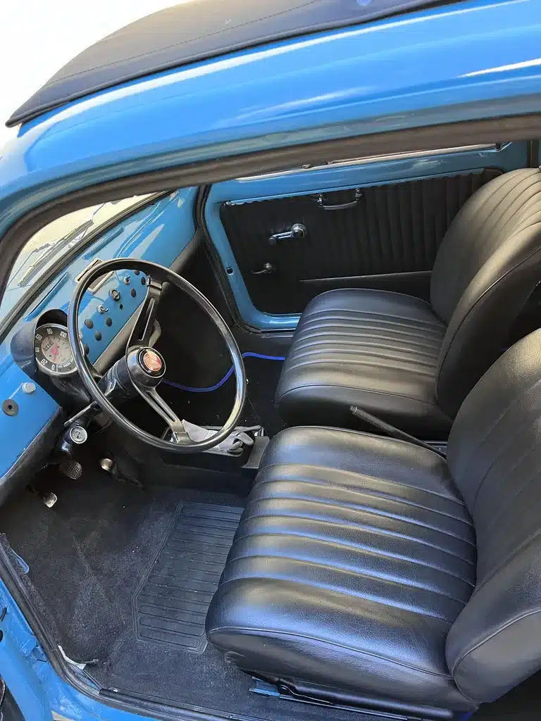 Blick in den Innenraum eines blauen Fiat 500L von 1968 mit schwarzen Vinylsitzen, Armaturenbrett und Faltdach.