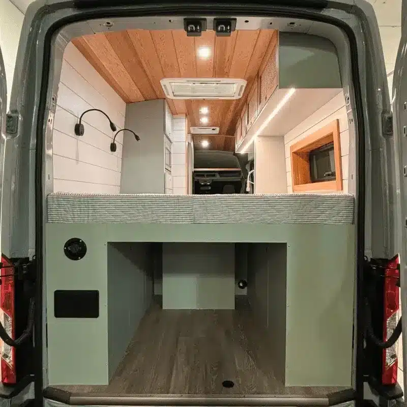 Der geöffnete Heckbereich eines Ford Transit Bon Echo Campervan, sichtbar sind das Bett, Stauraum und der modern ausgebaute Innenraum mit Holzdecke.