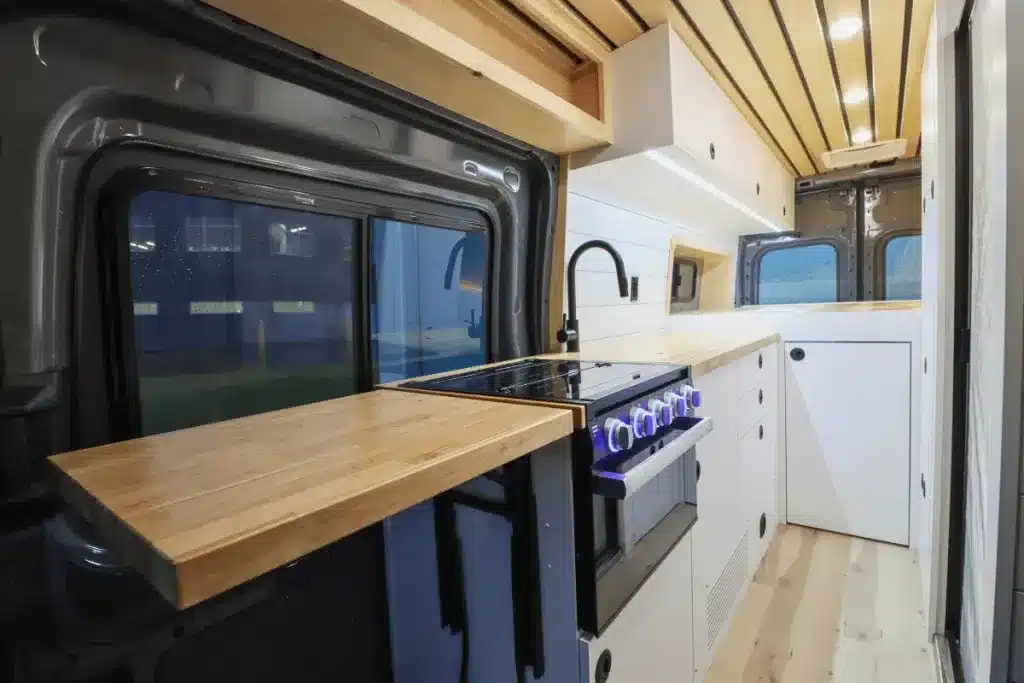 Innenansicht einer maßgefertigten Campervan-Küche im Ford Transit mit Holzarbeitsplatte, Induktionskochfeld, Gaskochfeld und modernen Stauraumlösungen.