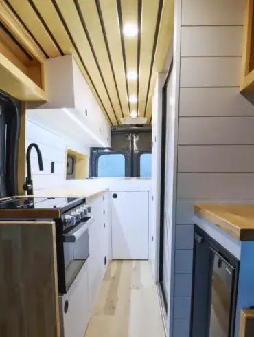 Innenansicht eines Campervans auf Ford Transit Basis mit moderner Bordküche, weißem Ausbau und Holzdetails.