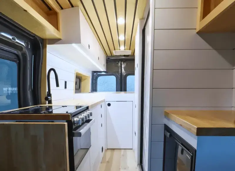 Innenansicht eines Campervans auf Ford Transit Basis mit moderner Bordküche, weißem Ausbau und Holzdetails.