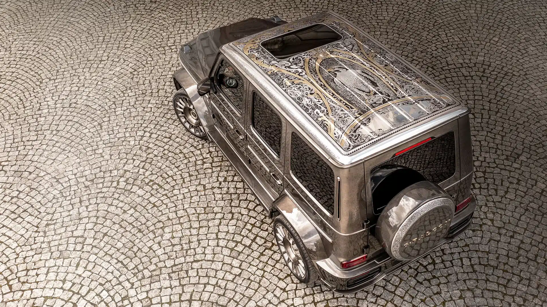Mercedes-Benz G 63 AMG G-Falcon One of One mit handgraviertem Falken-Relief auf dem Dach, diamantbesetzter Lackierung und auffälligen Felgen, fotografiert von schräg oben auf Kopfsteinpflaster.