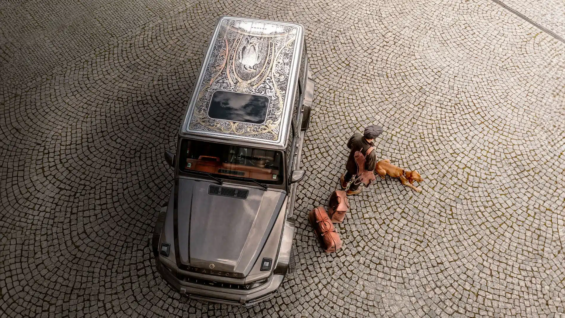 Mercedes-AMG G 63 G-FALCON One of One mit aufwendig graviertem Dach von oben fotografiert, auf Pflastersteinen, mit Person, Hund und Ledergepäck.