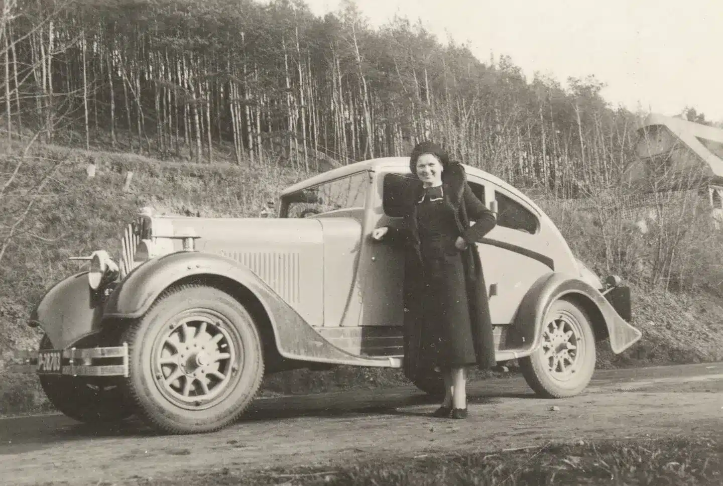 Schwarzweiß-Fotografie einer Frau mit einem historischen Škoda-Auto der 1930er-Jahre am Straßenrand vor einem Wald.