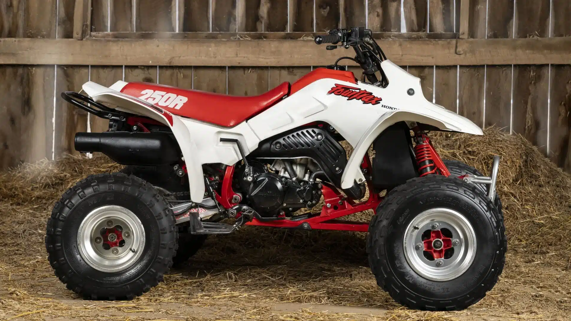 Seitenansicht einer weißen und roten Honda TRX250R Fourtrax von 1989 in einer Scheune mit Stroh am Boden