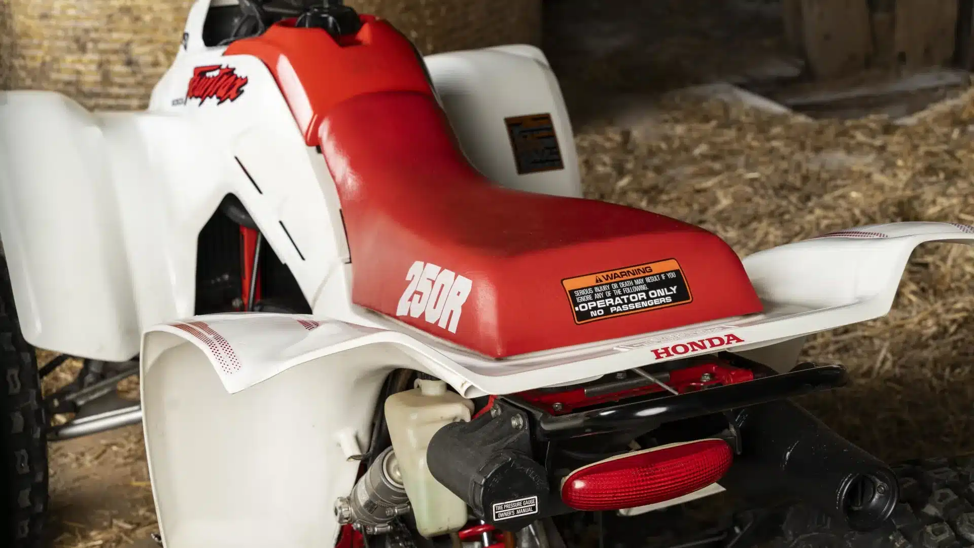 Nahaufnahme der roten Sitzbank mit 250R-Schriftzug und Heck der Honda TRX250R Fourtrax von 1989, aufgenommen in einer Garage.