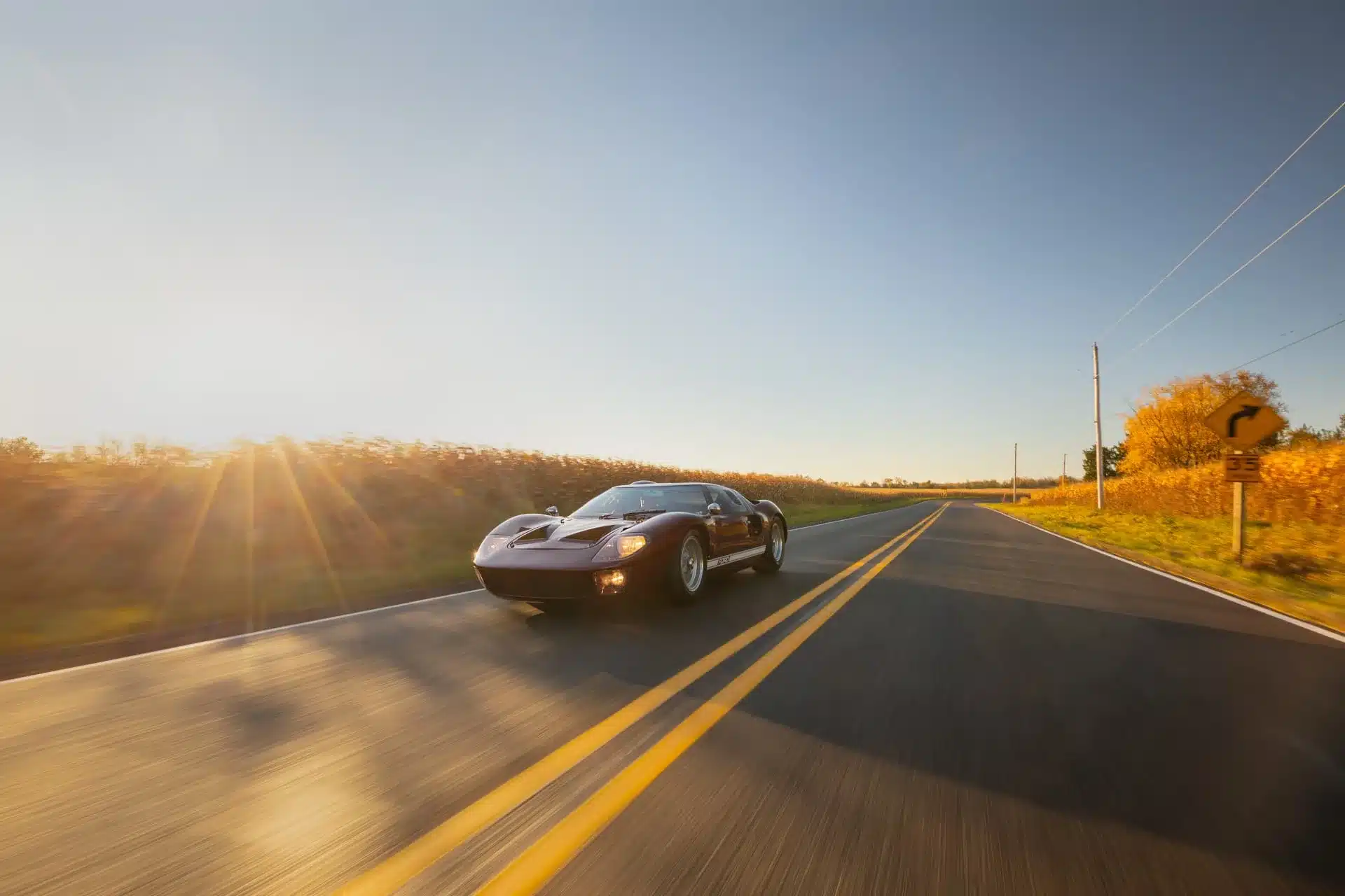 Ein 1965 Ford GT40 MkI in dunkler Lackierung fährt auf einer zweispurigen Landstraße bei Sonnenuntergang, flankiert von Feldern und Herbstbäumen.