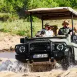 INEOS Grenadier Game Viewer fährt mit mehreren Personen über sandigen Untergrund und Wasser, typischer Safari-Einsatz.