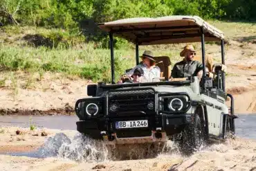 INEOS Grenadier Game Viewer fährt mit mehreren Personen über sandigen Untergrund und Wasser, typischer Safari-Einsatz.