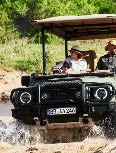 INEOS Grenadier Game Viewer fährt mit mehreren Personen über sandigen Untergrund und Wasser, typischer Safari-Einsatz.