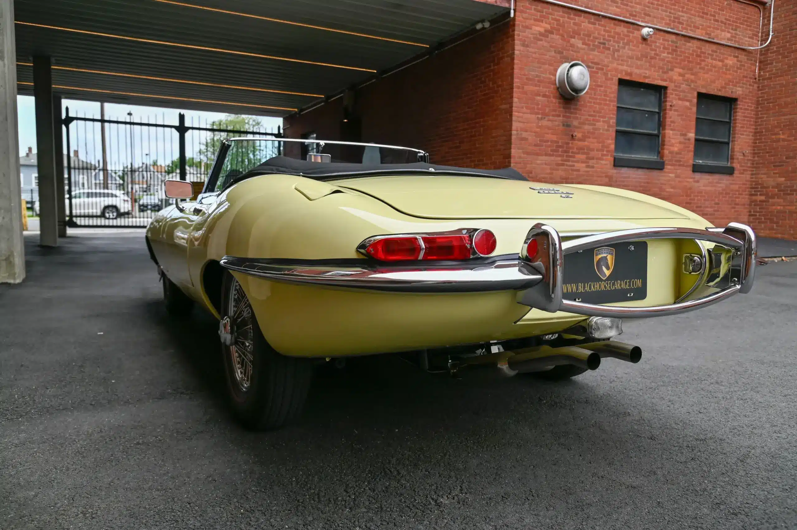 Rückansicht eines Jaguar E-Type Series 1 Roadsters, Farbe Pale Primrose, mit offenem Verdeck, Chromstoßstangen und roten Rückleuchten in einer Einfahrt.