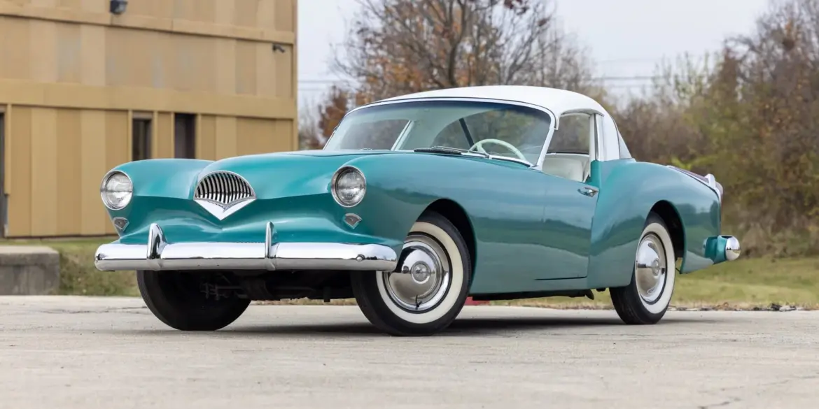Frontansicht eines türkis-weißen Kaiser Darrin 1954 Coupés auf einem Parkplatz, hervorgehoben durch Chromdetails und elegante Linienführung.