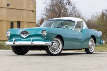Frontansicht eines türkis-weißen Kaiser Darrin 1954 Coupés auf einem Parkplatz, hervorgehoben durch Chromdetails und elegante Linienführung.