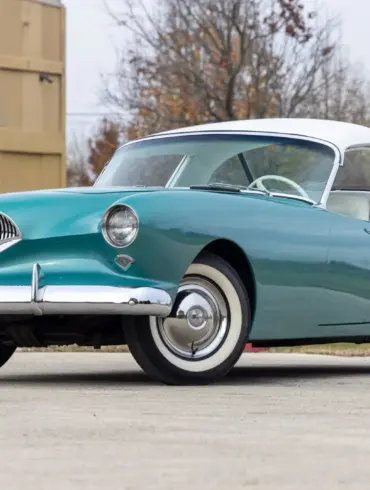 Frontansicht eines türkis-weißen Kaiser Darrin 1954 Coupés auf einem Parkplatz, hervorgehoben durch Chromdetails und elegante Linienführung.