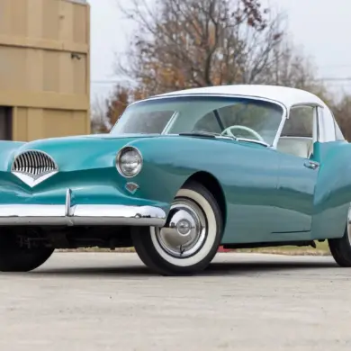 Frontansicht eines türkis-weißen Kaiser Darrin 1954 Coupés auf einem Parkplatz, hervorgehoben durch Chromdetails und elegante Linienführung.