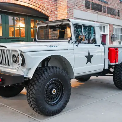 Komplett restaurierter Kaiser-Jeep M715 mit Offroad-Reifen, grauer Lackierung und modernen Ausstattungsdetails, abgestellt vor einem Backsteingebäude.