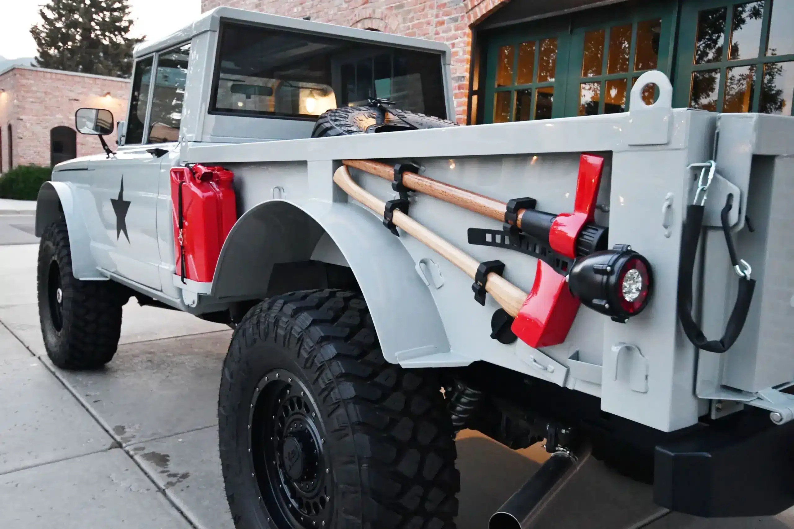 Seitliche Detailaufnahme eines grau lackierten Kaiser-Jeep M715 Restomods mit grobstolligen Offroad-Reifen, Reserverad, roten Reservekanistern und befestigten Werkzeugen an der Ladefläche.