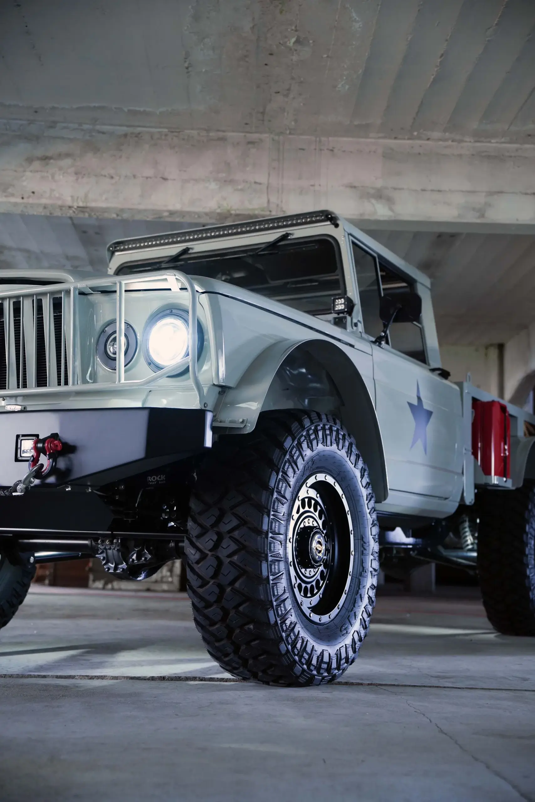 Kaiser-Jeep M715 Restomod mit großen Offroad-Reifen, LED-Scheinwerfern und Militärdekor in einer Parkgarage.