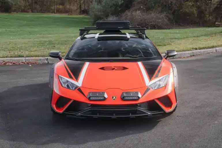 Frontansicht eines orangefarbenen Lamborghini Huracán Sterrato mit Offroad-Elementen auf einem asphaltierten Parkplatz.