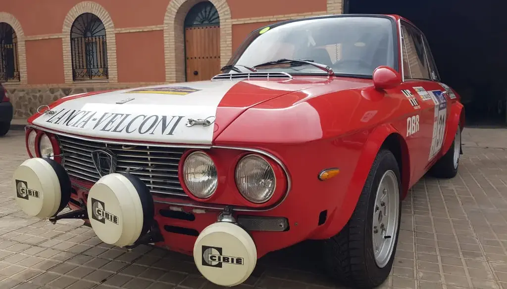 Lancia Fulvia HF 1600 Corsa Rallyefahrzeug, rot-weiß lackiert mit Zusatzscheinwerfern, Rallyeaufklebern und Startnummer vor einer Garage.