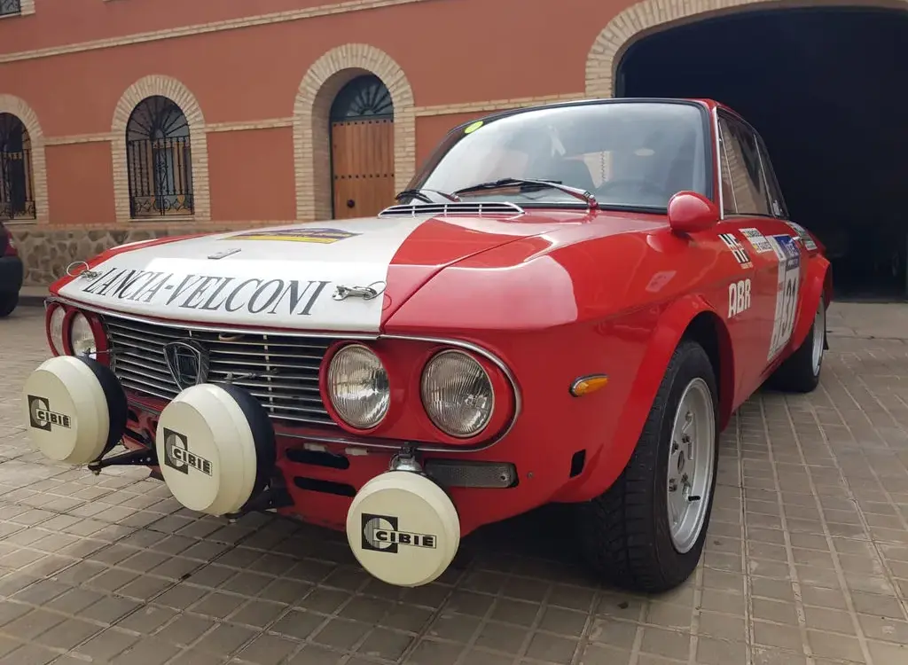 Lancia Fulvia HF 1600 Corsa Rallyefahrzeug, rot-weiß lackiert mit Zusatzscheinwerfern, Rallyeaufklebern und Startnummer vor einer Garage.