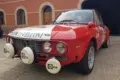 Lancia Fulvia HF 1600 Corsa Rallyefahrzeug, rot-weiß lackiert mit Zusatzscheinwerfern, Rallyeaufklebern und Startnummer vor einer Garage.