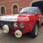 Lancia Fulvia HF 1600 Corsa Rallyefahrzeug, rot-weiß lackiert mit Zusatzscheinwerfern, Rallyeaufklebern und Startnummer vor einer Garage.