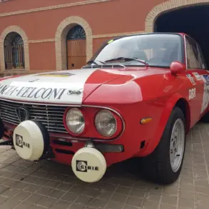 Lancia Fulvia HF 1600 Corsa Rallyefahrzeug, rot-weiß lackiert mit Zusatzscheinwerfern, Rallyeaufklebern und Startnummer vor einer Garage.