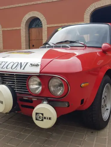 Lancia Fulvia HF 1600 Corsa Rallyefahrzeug, rot-weiß lackiert mit Zusatzscheinwerfern, Rallyeaufklebern und Startnummer vor einer Garage.