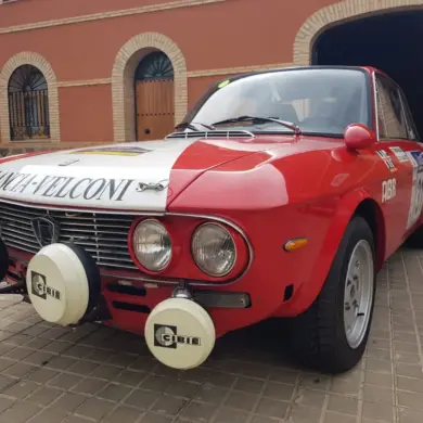 Lancia Fulvia HF 1600 Corsa Rallyefahrzeug, rot-weiß lackiert mit Zusatzscheinwerfern, Rallyeaufklebern und Startnummer vor einer Garage.