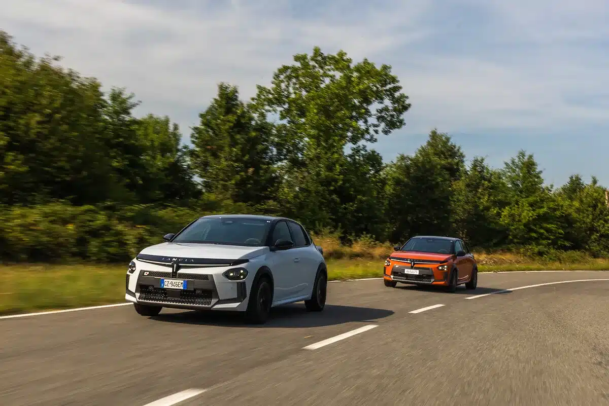 Zwei Lancia Ypsilon HF Modelle, in Weiß und Orange, fahren dynamisch auf einer Landstraße vor grünem Hintergrund
