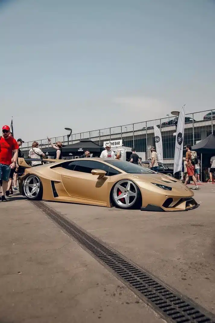 Goldener Liberty Walk Lamborghini Huracán LP610‑4 mit Widebody-Kit auf einem Eventgelände, tiefgelegt mit Rotiform Felgen und auffälligen Carbon-Details.
