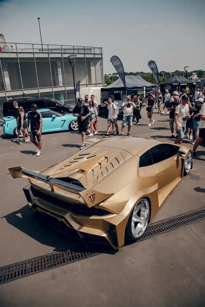 Liberty Walk LB‑Silhouette Works Lamborghini Huracán LP610‑4 in Gold mit Widebody-Kit und großem Heckflügel auf einer Veranstaltung, umgeben von Menschen.