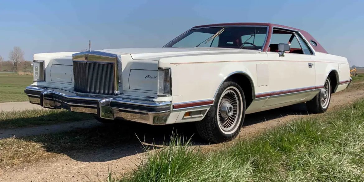 Weißer Lincoln Continental MK V Coupé, Baujahr 1977, mit rotem Vinyl-Dach, stehend auf einer ländlichen Straße, im Vordergrund Wiese und blauer Himmel.