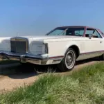 Weißer Lincoln Continental MK V Coupé, Baujahr 1977, mit rotem Vinyl-Dach, stehend auf einer ländlichen Straße, im Vordergrund Wiese und blauer Himmel.