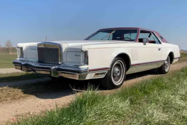 Weißer Lincoln Continental MK V Coupé, Baujahr 1977, mit rotem Vinyl-Dach, stehend auf einer ländlichen Straße, im Vordergrund Wiese und blauer Himmel.