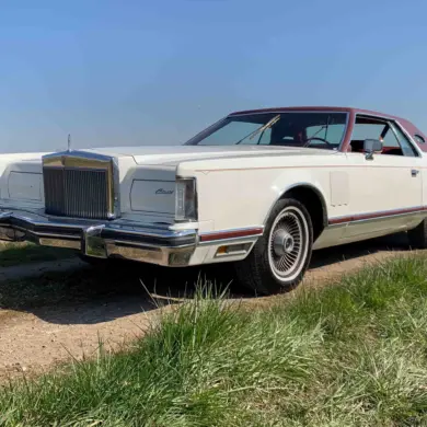 Weißer Lincoln Continental MK V Coupé, Baujahr 1977, mit rotem Vinyl-Dach, stehend auf einer ländlichen Straße, im Vordergrund Wiese und blauer Himmel.