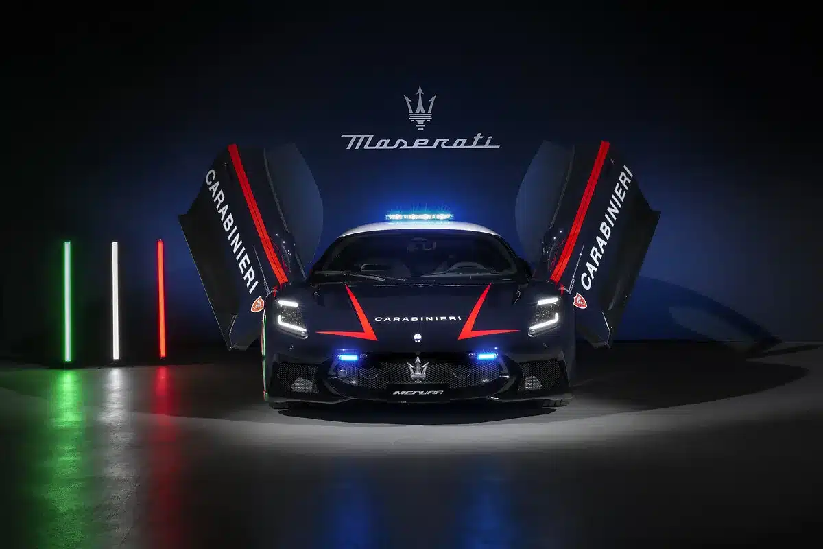 Frontansicht eines Maserati MCPURA in Carabinieri-Lackierung mit geöffneten Flügeltüren, eingeschaltetem Blaulicht und italienischer Flagge im Hintergrund.