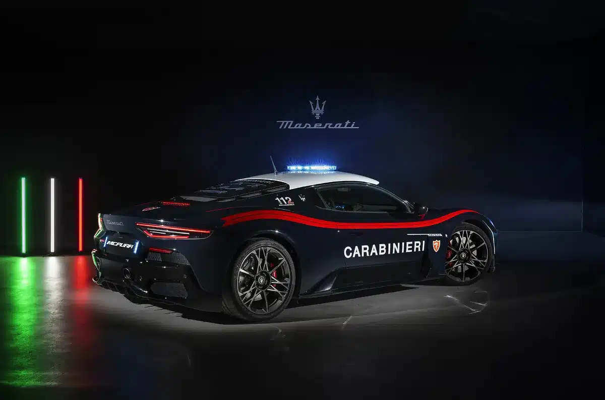 Maserati MCPURA in Carabinieri-Lackierung mit Blaulicht, seitliche Ansicht in dunkler Halle neben italienischer Flagge.