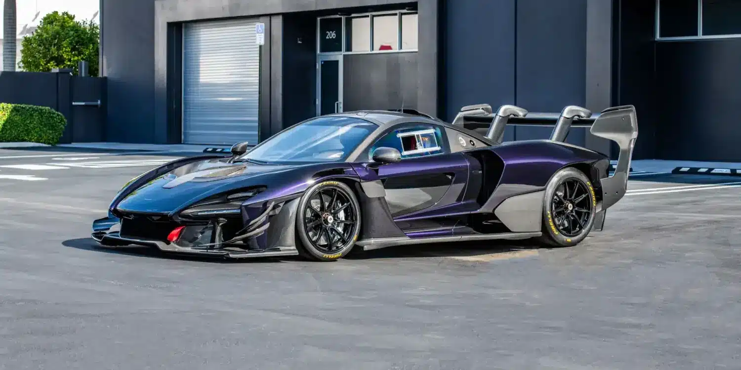 Seitliche Ansicht eines McLaren Senna GTR in dunklem Violett mit markantem Heckflügel und Rennsportdetails, geparkt vor einem modernen Industriegebäude.