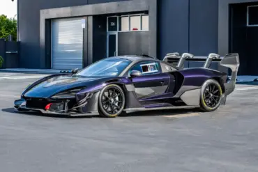 Seitliche Ansicht eines McLaren Senna GTR in dunklem Violett mit markantem Heckflügel und Rennsportdetails, geparkt vor einem modernen Industriegebäude.